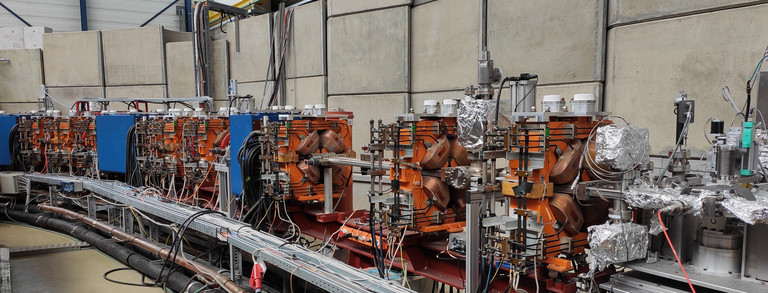 Strahlkammer mit Magneten Vom rechten Bildrand nach links verläuft ein Teil der Vakuumkammer des Speicherrings. Sie ist von blauen und orangen Magnten umschlossen. Im Hintergrund ist die Betonmauer zu erkennen.