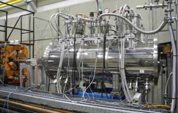 Supraleitender Wiggler Der supraleitende Wiggler ist im Speicherring installiert. Man sieht einen metallischen Zylinder mit zahlreichen Anschlussleitungen. Links im Bild ist ein orangefarbender Quadrupolmagnet.